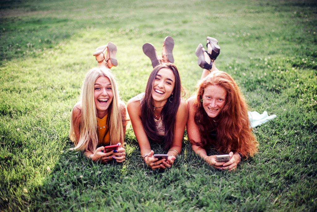 three young friends with long hair sitting on grass in skirts and sleeveless tops on a sunny summer day